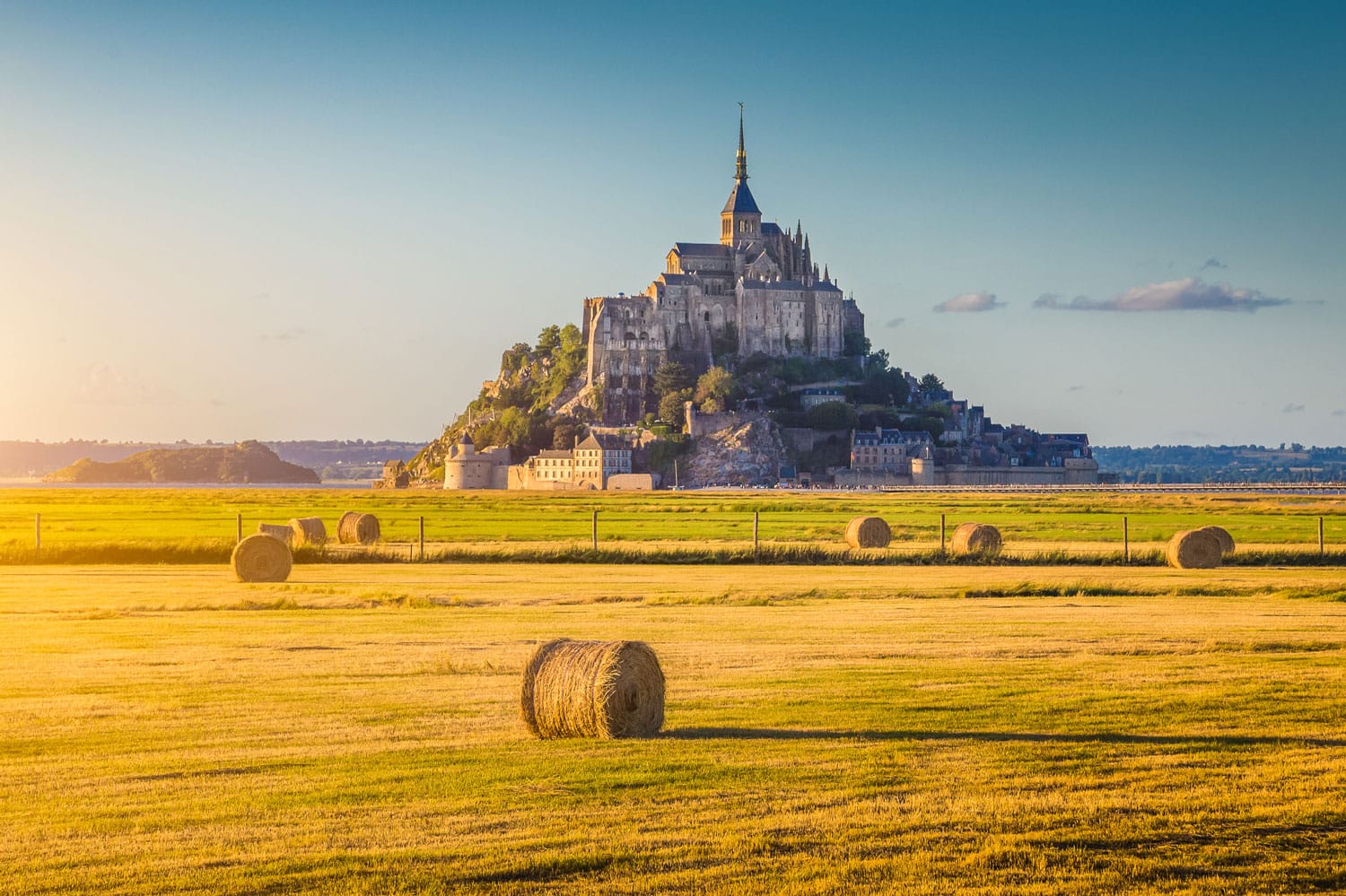 Beautiful view of famous historic Le Mont Saint-Michel tidal island in beautiful golden evening light at sunset in summer with hay bales on empty fields, Normandy, northern France