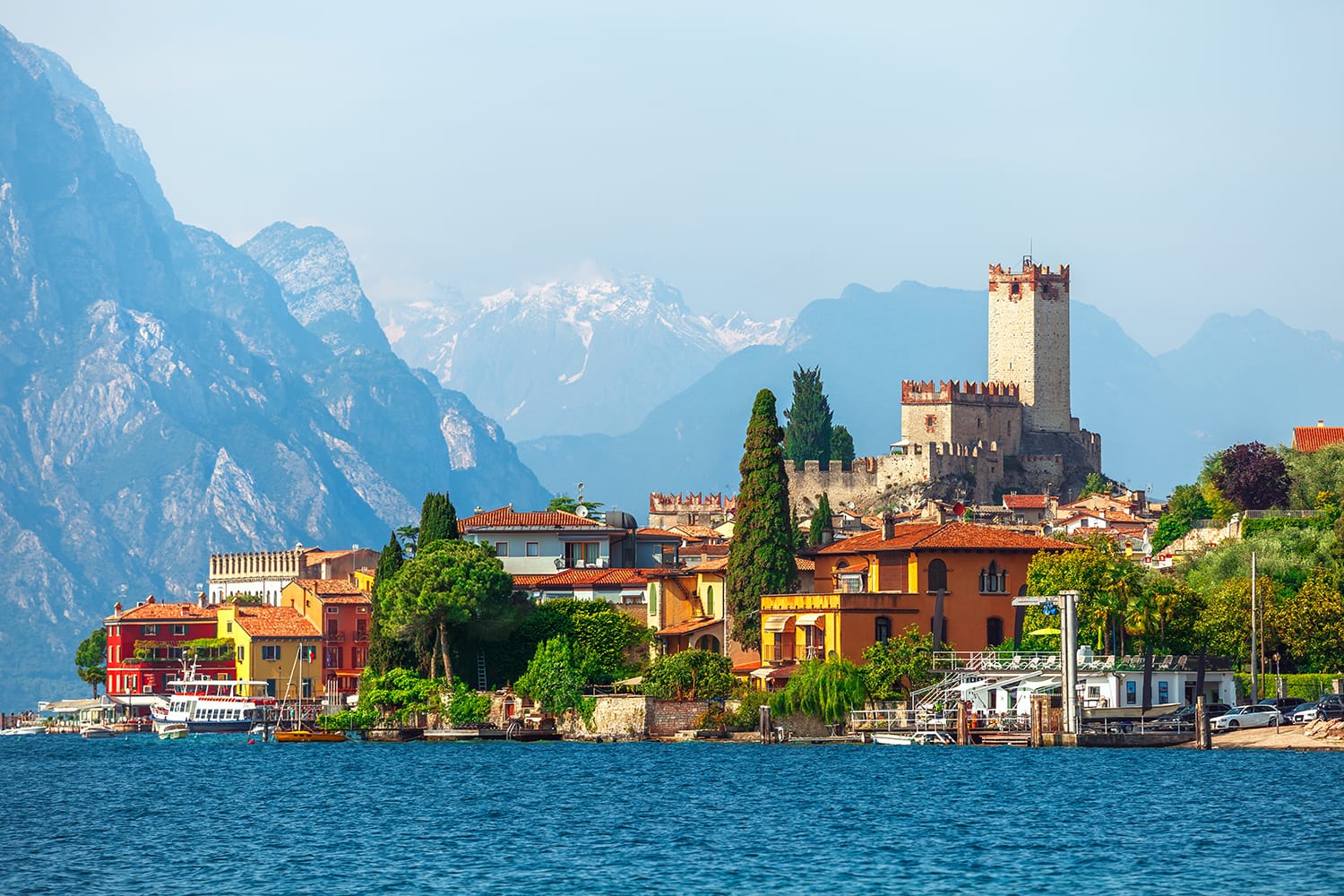 Malcesine town. Ancient tower and fortress in old town malcesine at Garda Lake, Veneto region, Italy. High snowbound top mountains on background summer landscape with colorful houses of Malcesine bank