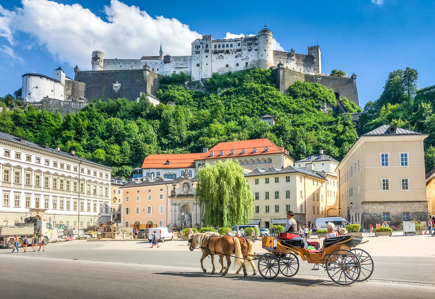 Beautiful panoramic view of the historic city of Salzburg with traditonal horse-drawn Fiaker carriage and famous Hohensalzburg Fortress on a hill on a sunny day with blue sky and clouds in summer