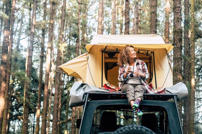 Happy hipster traveler people enjoying the alternative vacation - car with tent on the roof and beautiful young woman sit down outside looking at the forest around - outdoor activity
