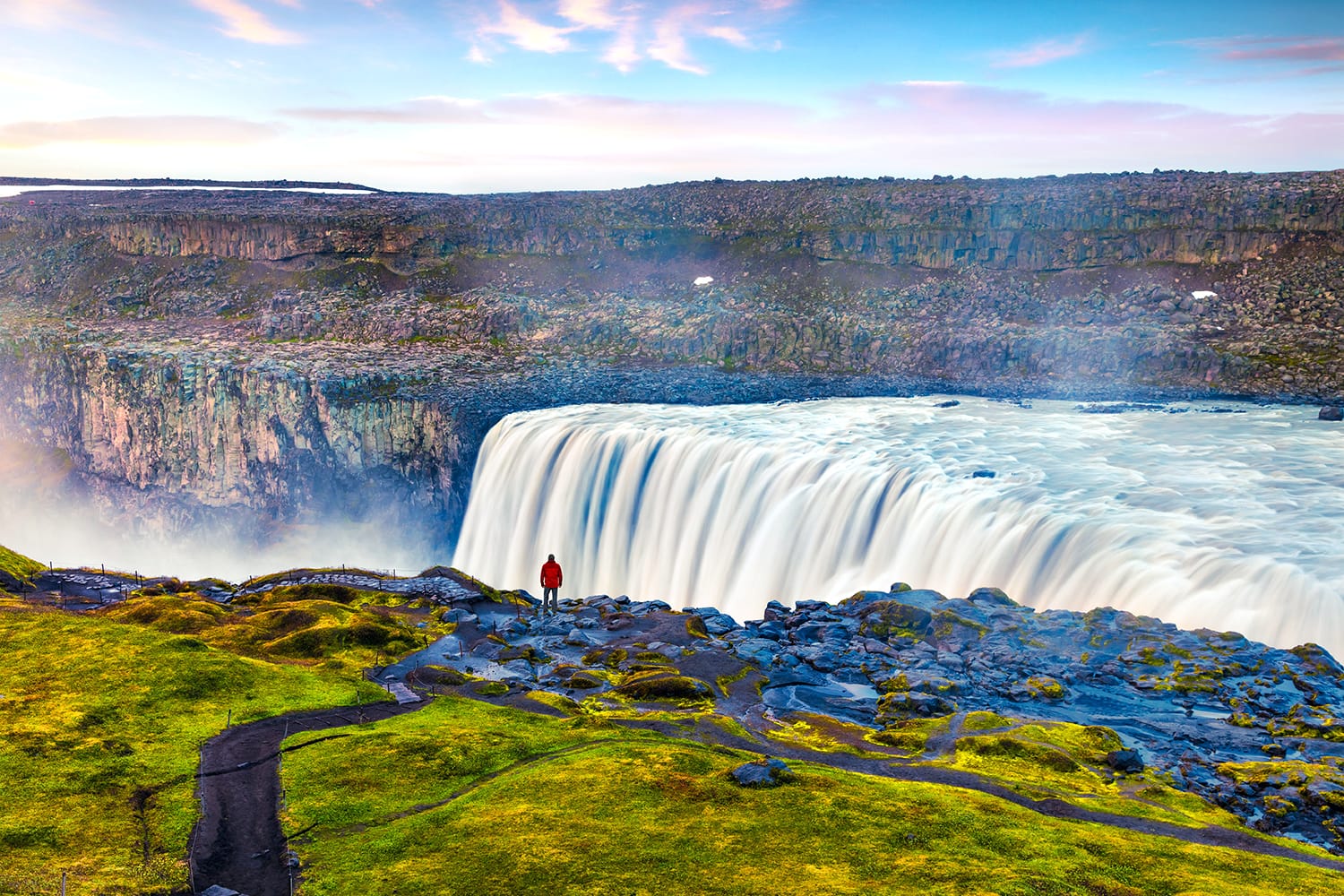 Tourist admiring view of falling water of the most powerful waterfall in Europe - Dettifoss. Colorful summer sunrise in Jokulsargljufur National Park, Iceland.