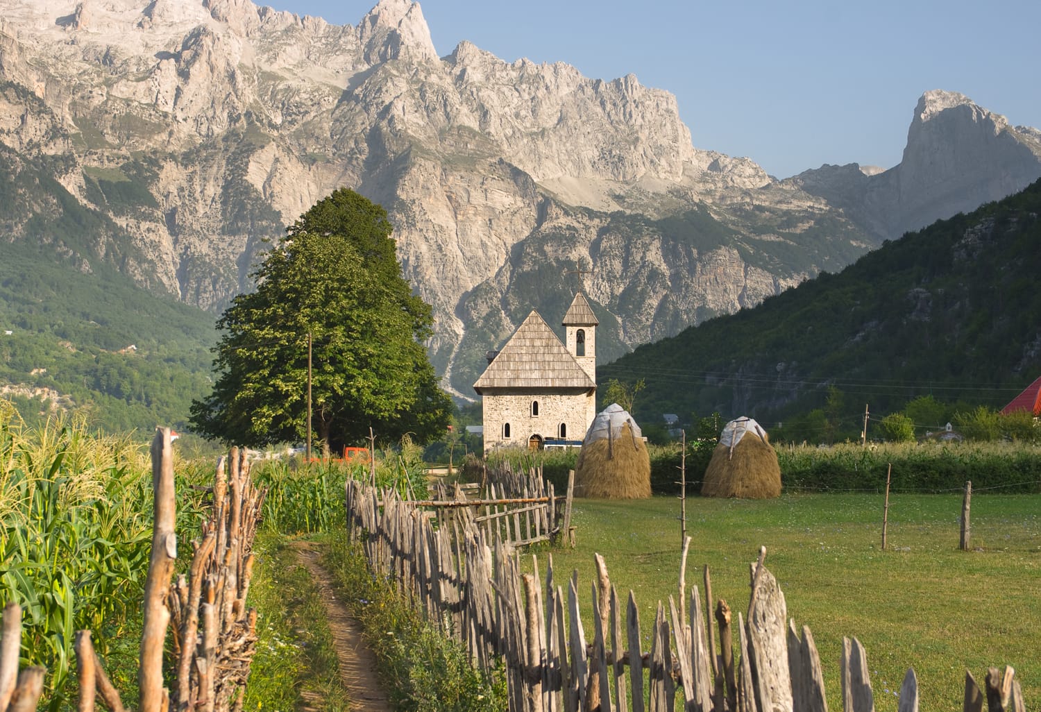 a path with wooden fence leads the catholic church in the Theth Valley, on background the mountains of Albanian Alps