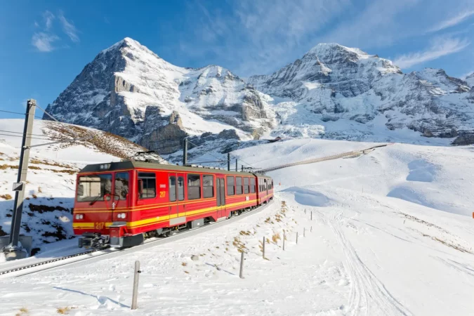 On a sunny winter day, tourists ride on a cogwheel train from Jungfraujoch (Top of Europe) to Kleine Scheidegg on the snowy hillside with Eiger and Monch in background, in Berner Oberland, Switzerland