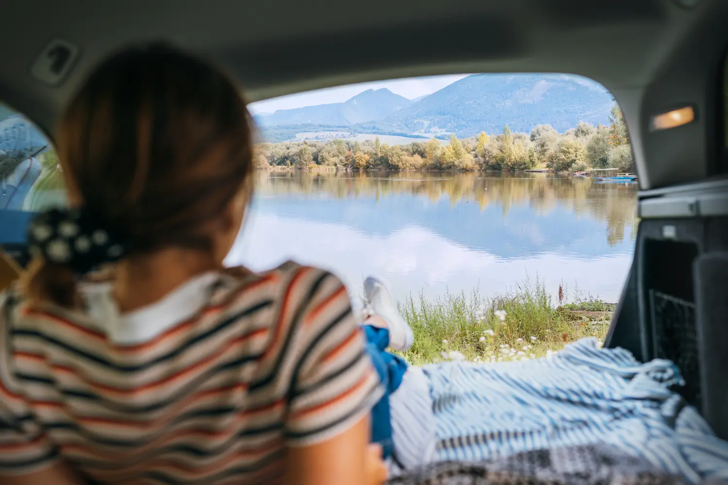 Woman lying in the cozy car trunk and enjoying the mountain lake view in the morning