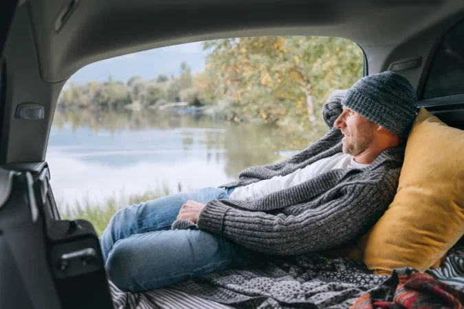 Smiling middle-aged man dressed in warm knitted clothes and jeans lying in the cozy car trunk and enjoying the mountain lake view.